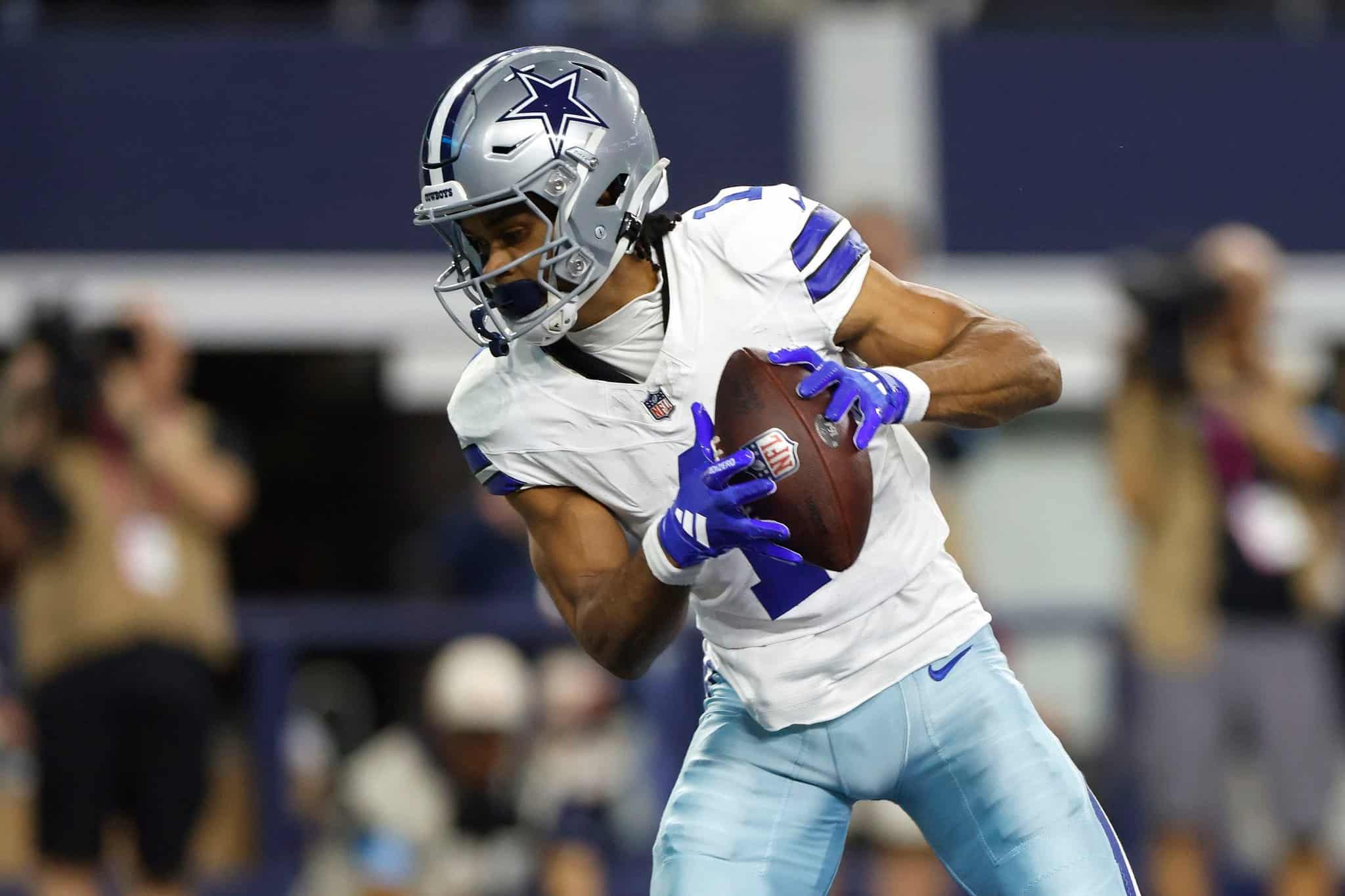 Dallas Cowboys player holding football during game, wearing helmet and white uniform, focused on play, surrounded by blurred crowd, inside the stadium during NFL game.