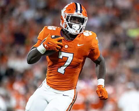 High school football player in orange uniform and helmet running with football on the field during game.
