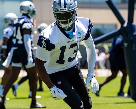 Fast-paced Dallas Cowboys football player running drills during practice at training camp. The player is wearing a white jersey with number 13, black pants, and a silver helmet, training outdoors on a sunny day.