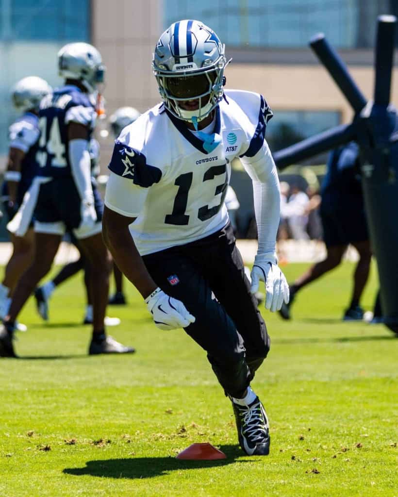 Fast-paced Dallas Cowboys football player running drills during practice at training camp. The player is wearing a white jersey with number 13, black pants, and a silver helmet, training outdoors on a sunny day.