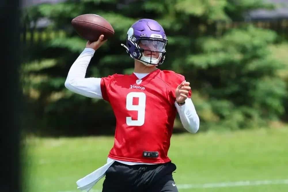 Quarterback practicing football throw during a game, wearing a red jersey with the number 9 and a purple helmet, on a lush green field.