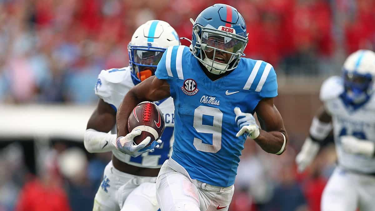 Mid-game football action featuring a player from Ole Miss rushing with the ball during a college football game. The player is wearing a blue uniform with the number 9, with opposing team players in white and blue uniforms in pursuit. The background shows a blurred crowd and stadium atmosphere.