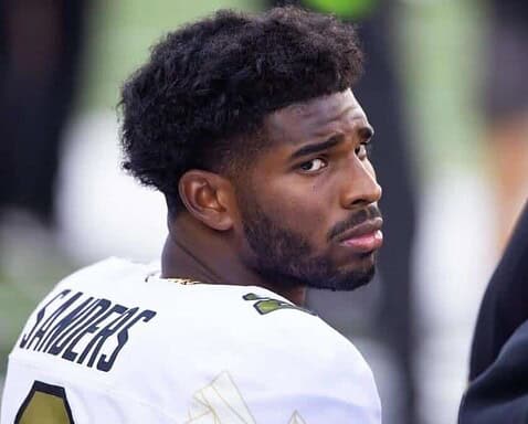 Young African American football player in a Dallas Cowboys jersey with "Shultz" on the back, looking back with a serious expression, at a game or practice, with a blurred crowd background.