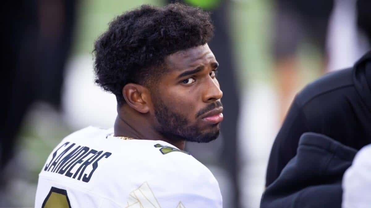 Young African American football player in a Dallas Cowboys jersey with "Shultz" on the back, looking back with a serious expression, at a game or practice, with a blurred crowd background.