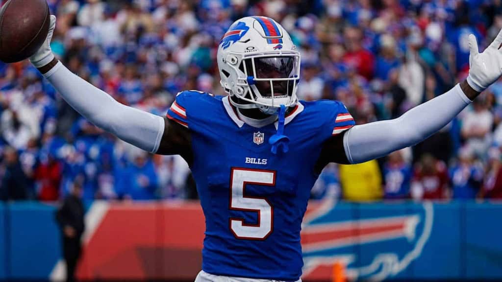 High-energy football player celebrating on the field in a Buffalo Bills uniform with arms outstretched during a game, with a crowd in the background.
