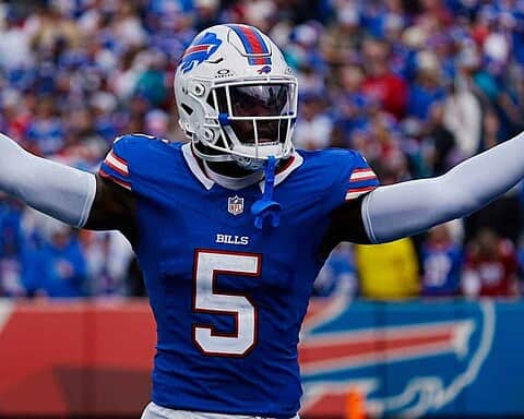 High-energy football player celebrating on the field in a Buffalo Bills uniform with arms outstretched during a game, with a crowd in the background.