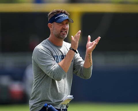 Coaching staff member guiding Dallas Cowboys during practice, wearing team apparel and a visor, discussing strategies on the field at a sunny training camp.