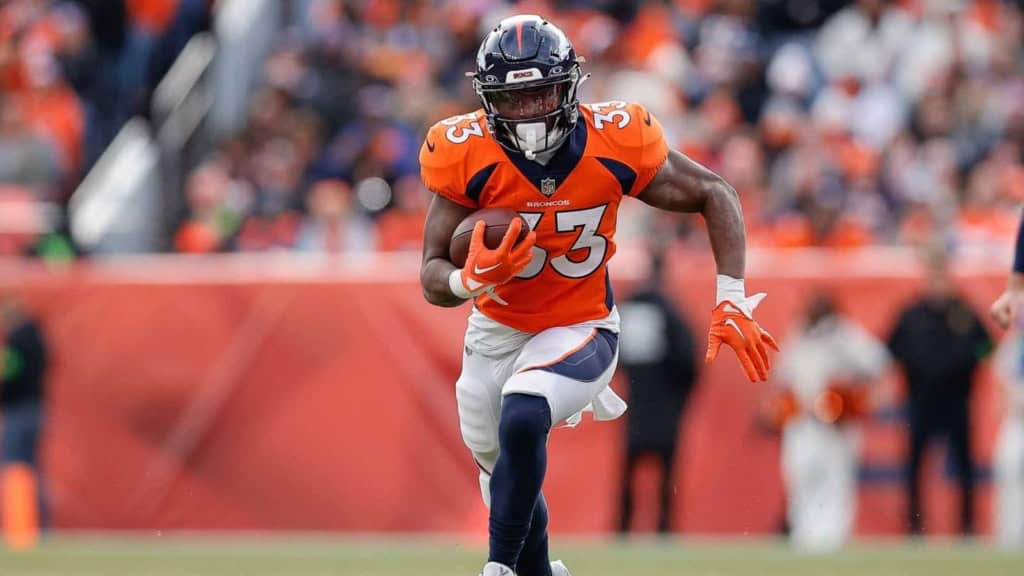 Steady running back for Denver Broncos with football, wearing orange jersey, during an NFL game in a stadium with a blurred crowd background.