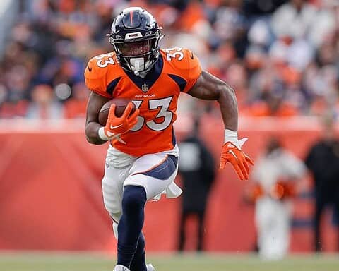 Steady running back for Denver Broncos with football, wearing orange jersey, during an NFL game in a stadium with a blurred crowd background.