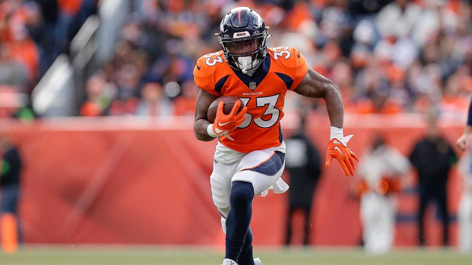 Steady running back for Denver Broncos with football, wearing orange jersey, during an NFL game in a stadium with a blurred crowd background.