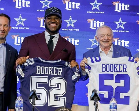 Dallas Cowboys player Trevon Diggs and owner Jerry Jones at a press conference or event, holding jerseys with the name "Booker" in front of a branded Dallas Cowboys backdrop. The event appears to celebrate or introduce new players or team members. The setting includes team branding, microphones, and water bottles on the table. The atmosphere is professional and celebratory, highlighting team loyalty and player introductions.