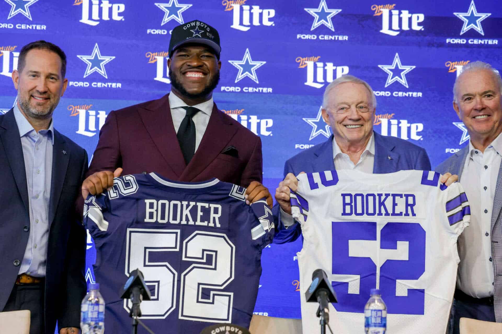 Dallas Cowboys player Trevon Diggs and owner Jerry Jones at a press conference or event, holding jerseys with the name "Booker" in front of a branded Dallas Cowboys backdrop. The event appears to celebrate or introduce new players or team members. The setting includes team branding, microphones, and water bottles on the table. The atmosphere is professional and celebratory, highlighting team loyalty and player introductions.