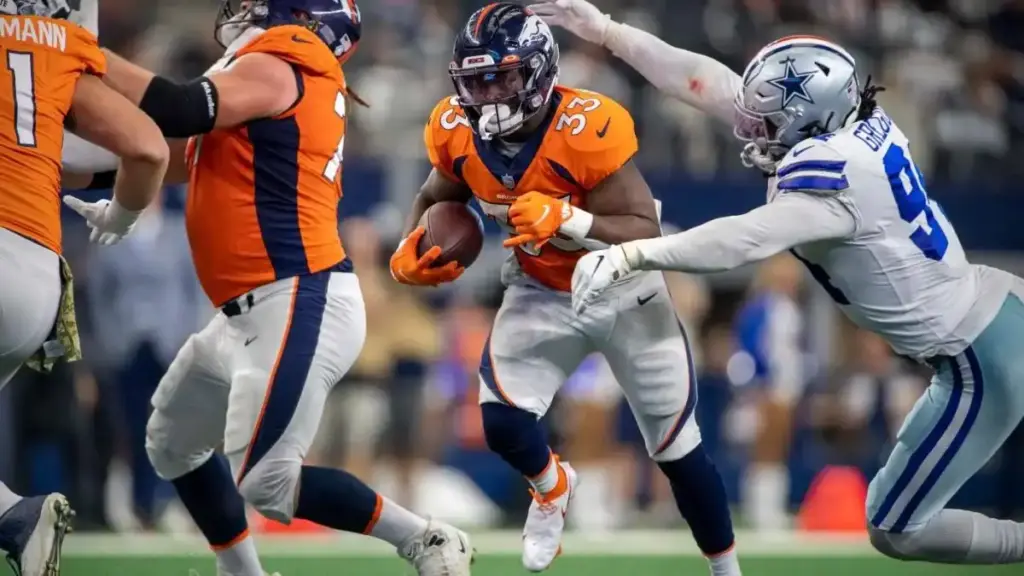 Broncos football player in orange uniform running with ball during game against Cowboys Dallas team.