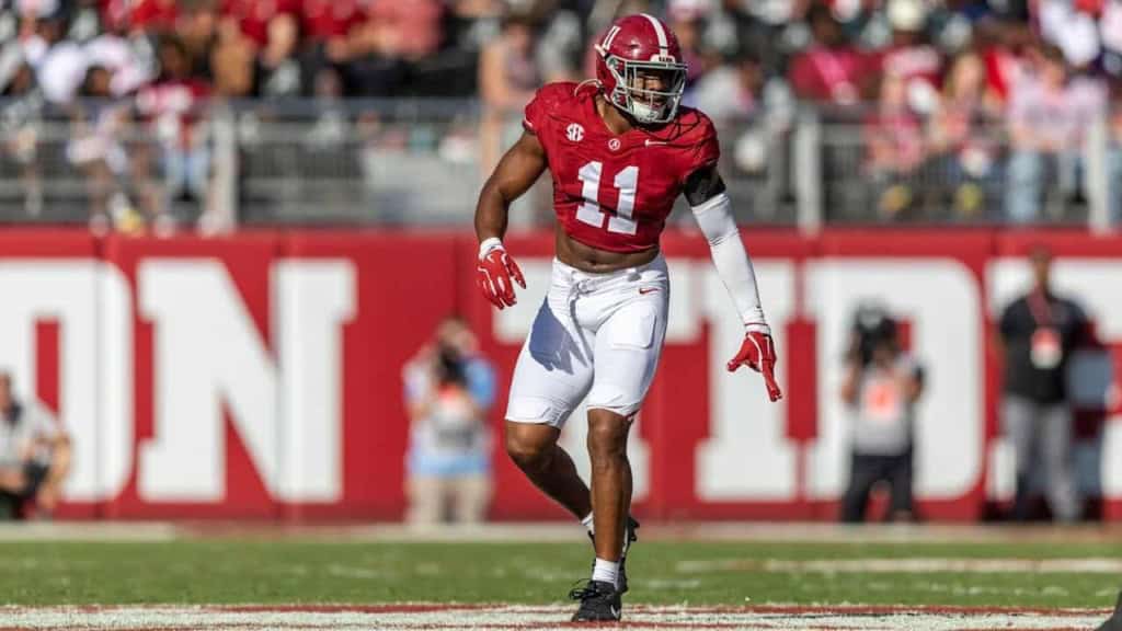 Alabama football player wearing number 11 in a game on the field, mid-play action, in full uniform including helmet, red jersey, and white pants, during a college football game.
