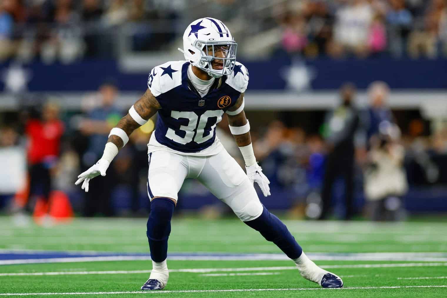 Dallas Cowboys football player in defensive stance during game at AT&T Stadium, focused on opponent, showcasing team uniform, helmet, and field environment.