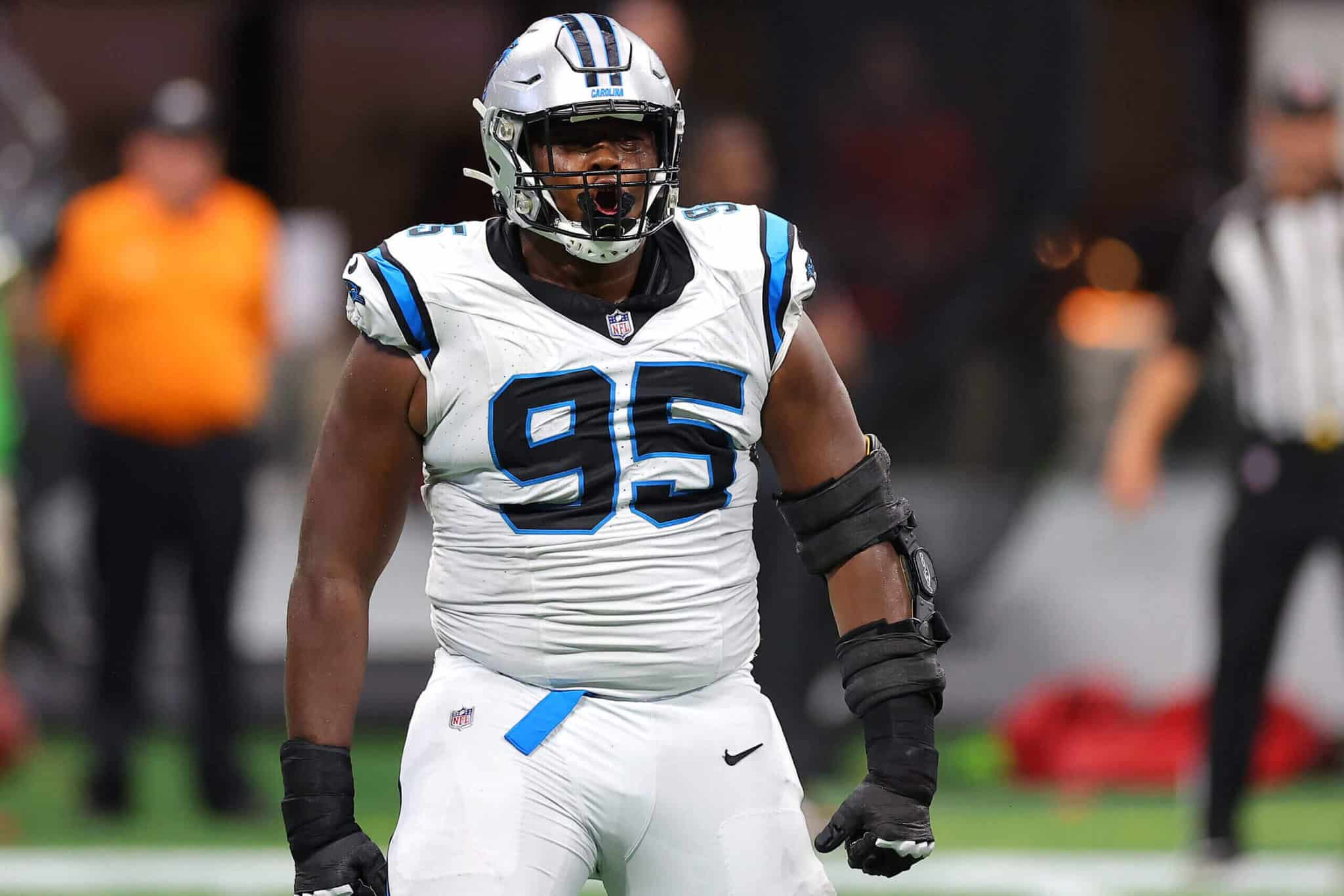 Aggressive football player in a white and blue NFL jersey number 95, wearing a helmet and protective arm gear, on the field celebrating a game moment.