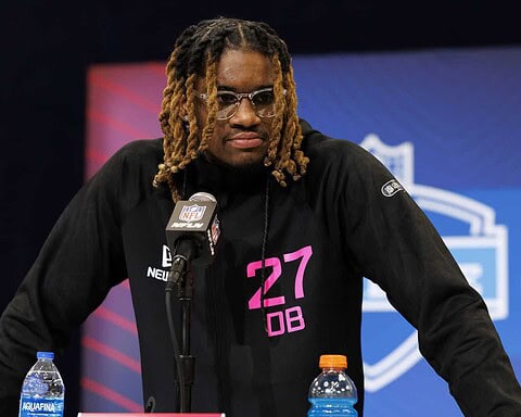 NFL player at press conference wearing black jacket with pink "27 DB" text, sitting at table with water bottles, NFL microphone, and NFL logo in background, inside The Star facility.