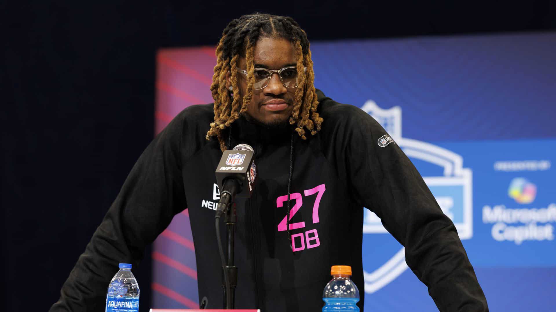 NFL player at press conference wearing black jacket with pink "27 DB" text, sitting at table with water bottles, NFL microphone, and NFL logo in background, inside The Star facility.