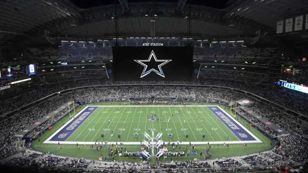Vast view of AT&T Stadium interior during a Dallas Cowboys football game, showing the field, large central scoreboard, and crowded stands filled with fans.