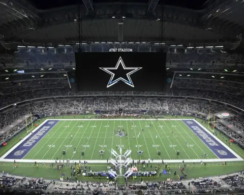 Vast view of AT&T Stadium interior during a Dallas Cowboys football game, showing the field, large central scoreboard, and crowded stands filled with fans.
