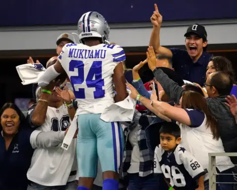 An NFL player from the Dallas Cowboys, wearing jersey number 24 with "MUKUAMU" on the back, celebrates with fans in the stadium, capturing excitement and team spirit at a football game.