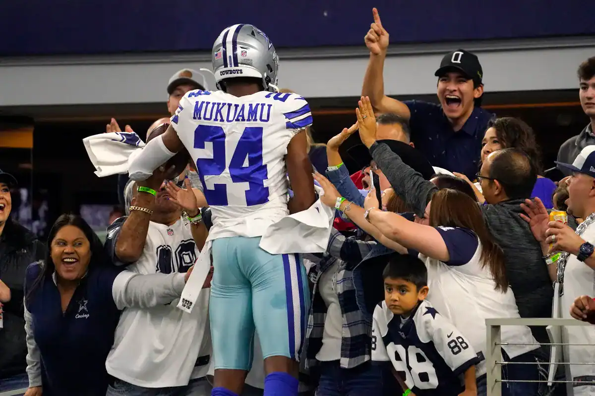 An NFL player from the Dallas Cowboys, wearing jersey number 24 with "MUKUAMU" on the back, celebrates with fans in the stadium, capturing excitement and team spirit at a football game.
