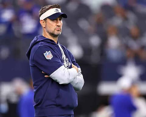 NFL coach standing with arms crossed during game at AT&T Stadium, wearing Dallas Cowboys hoodie and visor, focused on team strategy, inside the star.
