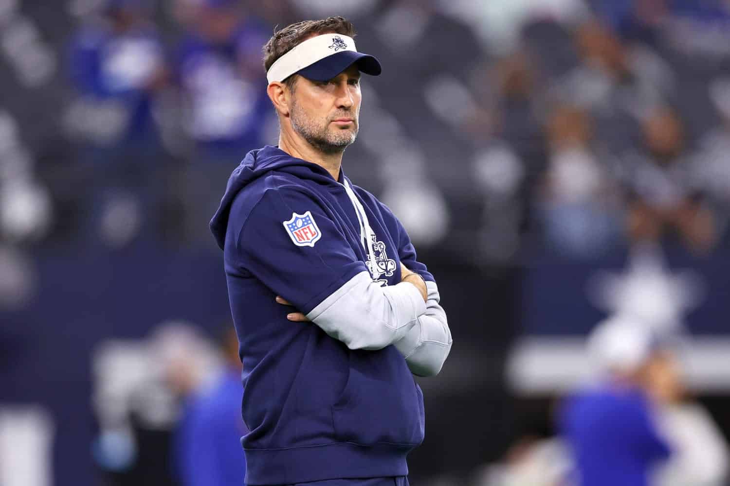 NFL coach standing with arms crossed during game at AT&T Stadium, wearing Dallas Cowboys hoodie and visor, focused on team strategy, inside the star.