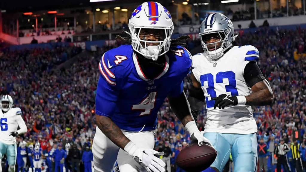 Eyebrow-raising NFL football game featuring Buffalo Bills player in blue jersey with number 4 catching a touchdown pass, and Dallas Cowboys player in white jersey with number 13 in close defense during a packed stadium.