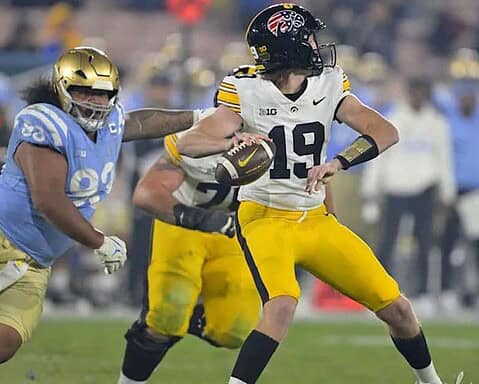 Fast-paced college football game featuring a University of Michigan quarterback running with the football against a rival team defender, with spectators in the background.