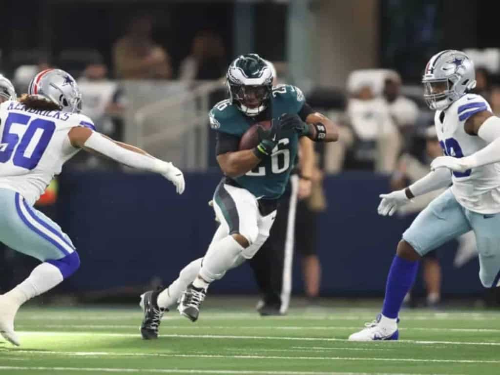 Busy football game action featuring a running back in a dark jersey holding the football while trying to evade defenders from the opposing team dressed in white uniforms.