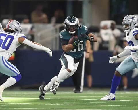 Busy football game action featuring a running back in a dark jersey holding the football while trying to evade defenders from the opposing team dressed in white uniforms.