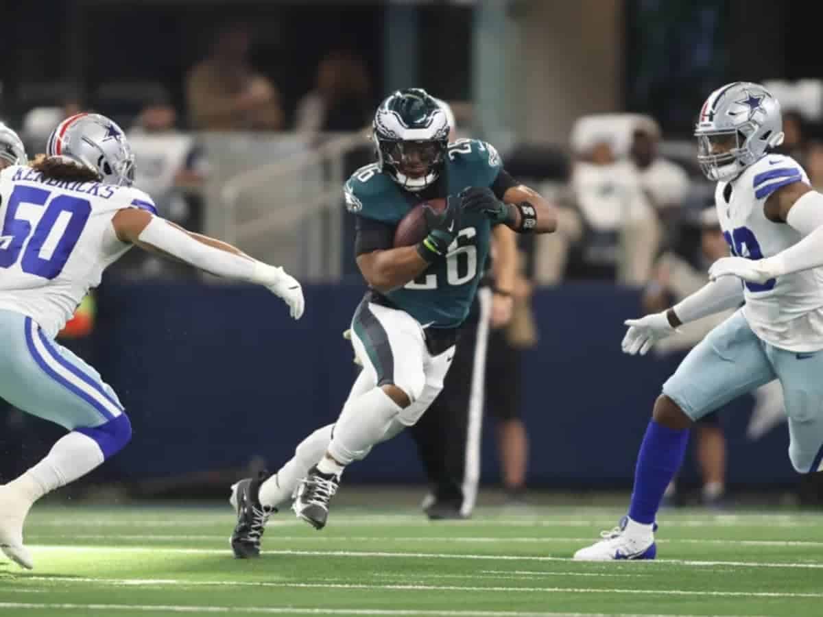 Busy football game action featuring a running back in a dark jersey holding the football while trying to evade defenders from the opposing team dressed in white uniforms.