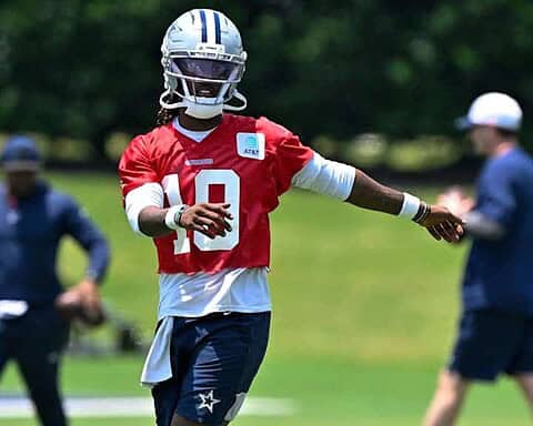 - Wide receiver Dak Prescott practicing during Dallas Cowboys training camp on a sunny day, wearing a red jersey and football helmet on lush green field.
