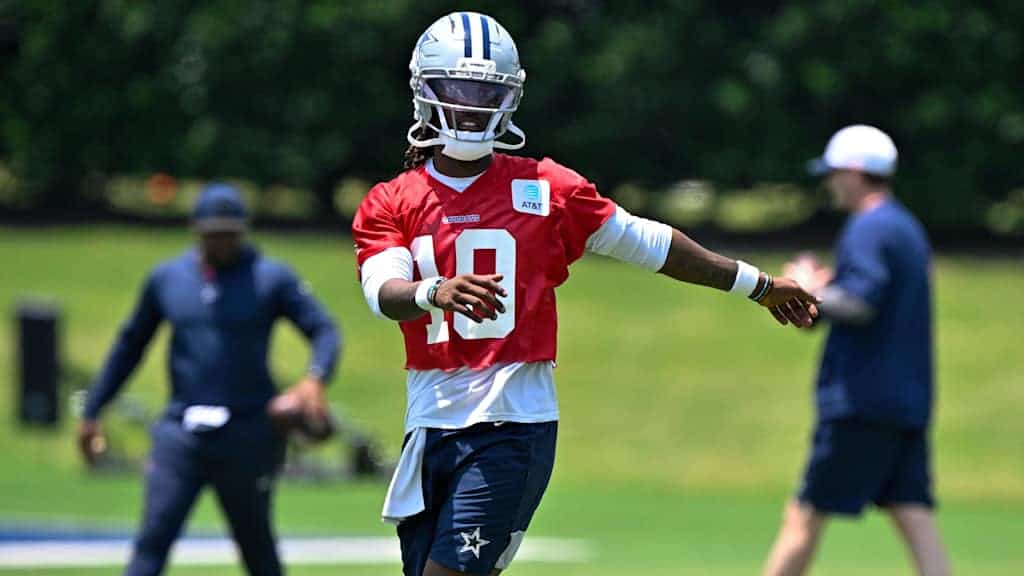 - Wide receiver Dak Prescott practicing during Dallas Cowboys training camp on a sunny day, wearing a red jersey and football helmet on lush green field.