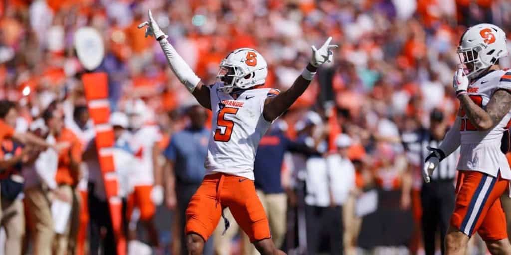 Syracuse football player celebrating with arms raised during game on the sidelines, wearing orange and white uniform.