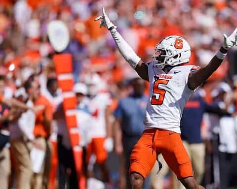 Syracuse football player celebrating with arms raised during game on the sidelines, wearing orange and white uniform.