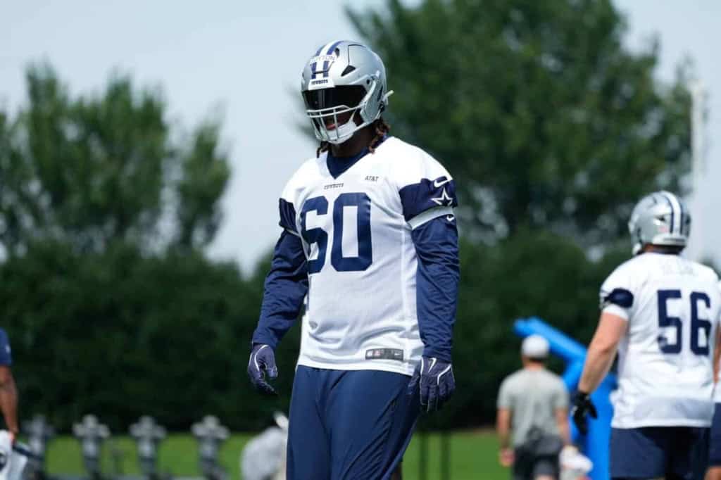 Dallas Cowboys football player during practice, wearing team uniform and helmet, on the field with green trees in the background.