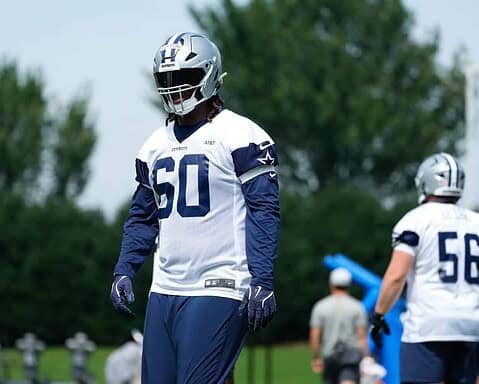 Dallas Cowboys football player during practice, wearing team uniform and helmet, on the field with green trees in the background.