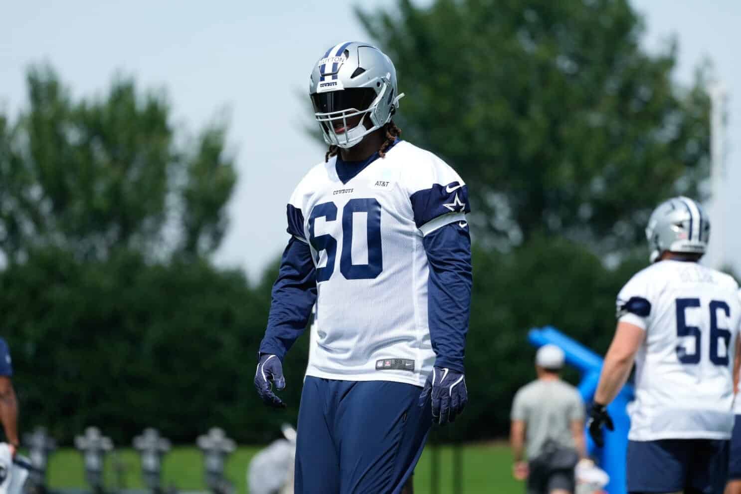 Dallas Cowboys football player during practice, wearing team uniform and helmet, on the field with green trees in the background.