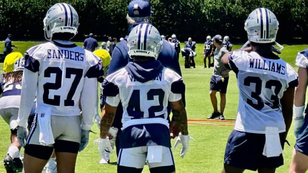 High school football players wearing Dallas Cowboys jerseys during practice or game on a sunny day, with coaches and players in the background.