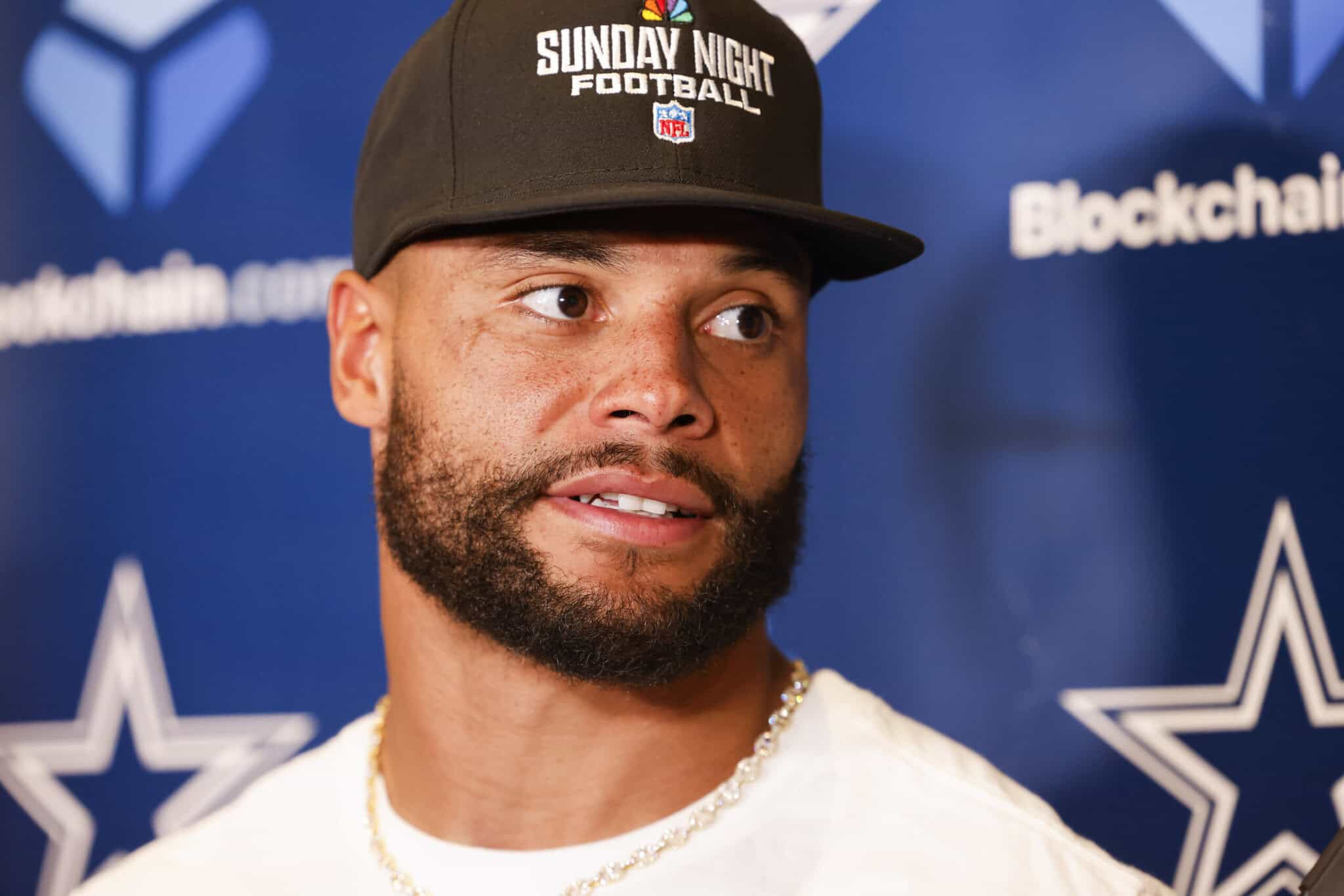 NFL player wearing a black hat with "Sunday Night Football" logo and chains, speaking at a press conference, with Dallas Cowboys branding in the background.