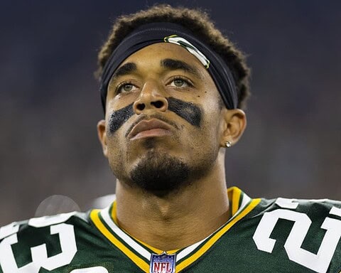 Focused Green Bay Packers football player wearing jersey with black eye paint, headband, and intense expression during game.