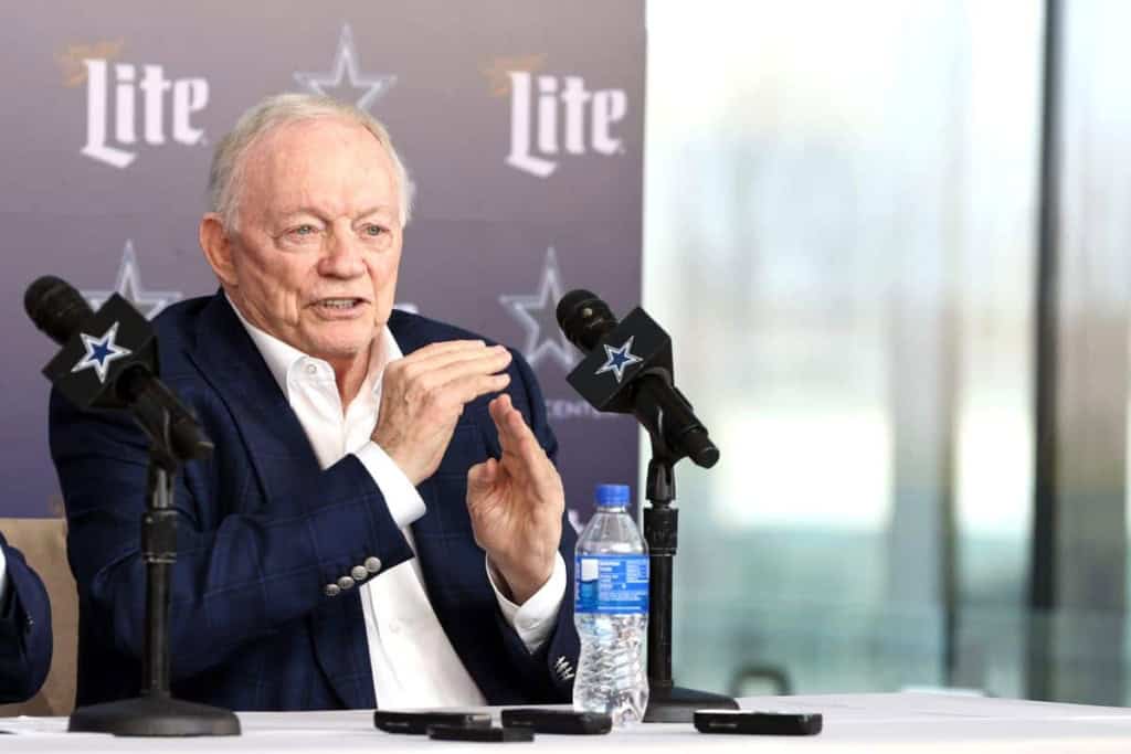 Veteran man speaking during press conference at Inside The Star, Dallas Cowboys NFL team, with team branding and microphones; sports management and football event.
