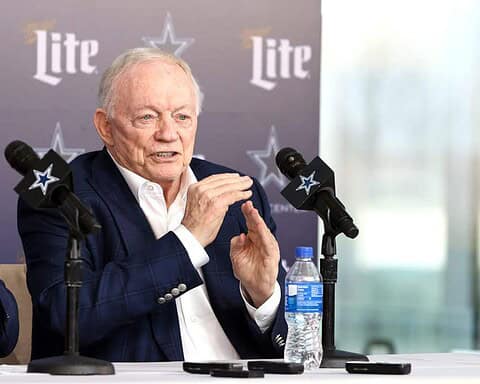 Veteran man speaking during press conference at Inside The Star, Dallas Cowboys NFL team, with team branding and microphones; sports management and football event.