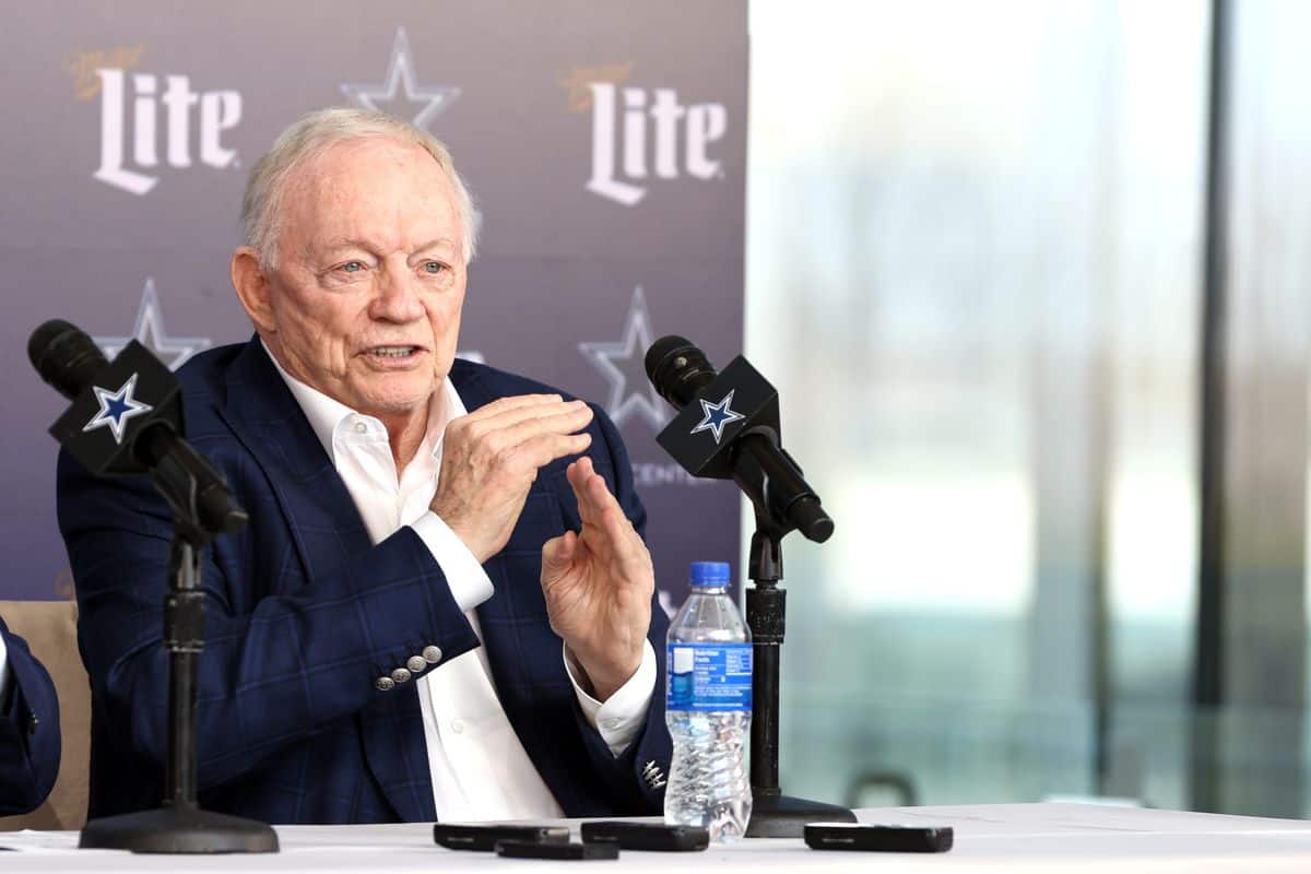 Veteran man speaking during press conference at Inside The Star, Dallas Cowboys NFL team, with team branding and microphones; sports management and football event.