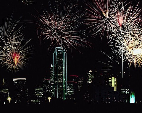 Vibrant fireworks display illuminating the Dallas skyline at night, featuring the Bank of America Plaza and other skyscrapers, celebrating with colorful explosions in the dark sky.
