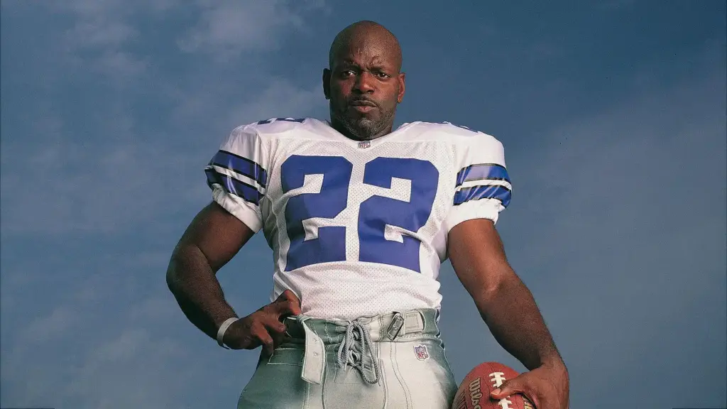 Pro NFL football player in a Dallas Cowboys jersey holding a football, standing outdoors against a clear blue sky, showcasing athlete strength and game readiness.