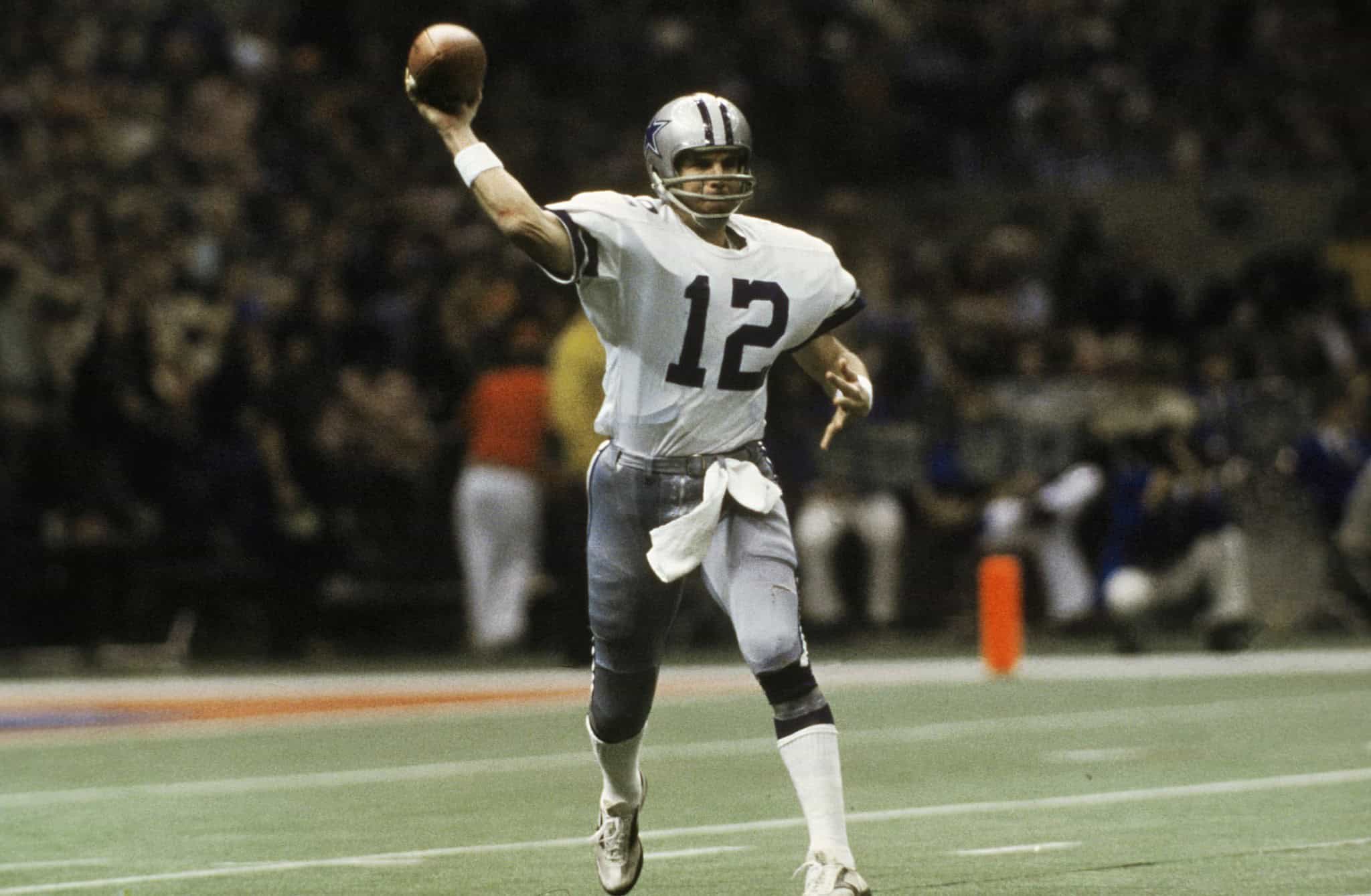 American football quarterback wearing Dallas Cowboys jersey number 12, preparing to throw a pass during a game, with a blurred crowd in the background.