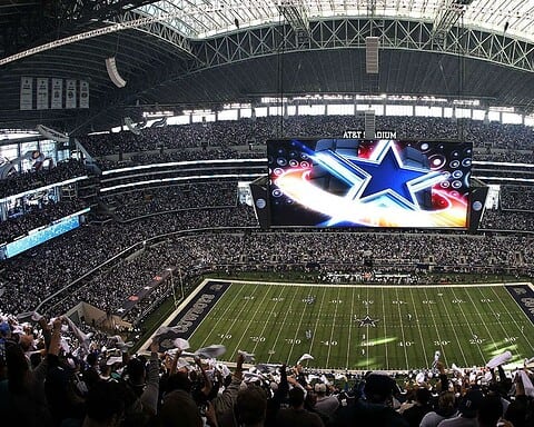 Dallas Cowboys stadium interior during a game, filled with cheering fans and a large digital screen displaying the Dallas Cowboys star logo, showcasing NFL football atmosphere.